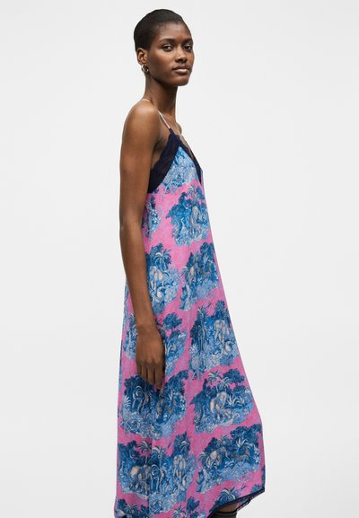 Γυναίκα που φοράει μακρύ ροζ και μπλε φόρεμα με λεπτές τιράντες και μαύρη δαντέλα, στέκεται προφίλ σε λευκό φόντο.