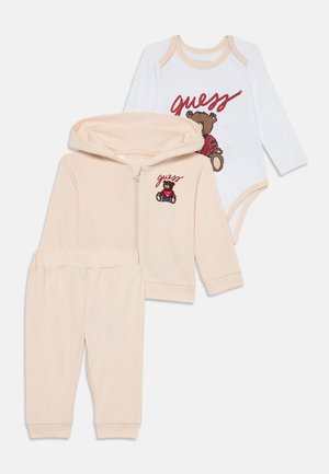 Tre-delt babyantrekk som inkluderer en beige hettegenser, matchende bukser og en hvit body med bjørnegrafikk og "guess"-tekst. Mykt bomullsmateriale.