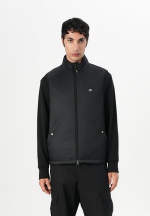 Black padded vest with a high collar, front zip closure, side pockets, and logo detail on the chest, worn over a long-sleeve black shirt.