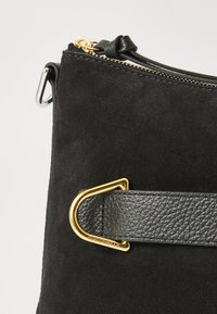 Borsa a tracolla in suede nera con una tracolla in pelle nera testurizzata e un accento in metallo dorato. Il design include una chiusura con zip nella parte superiore.