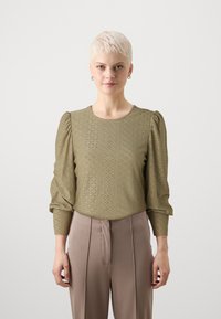 Olive green blouse with textured fabric featuring eyelet patterns, round neckline, and puffed long sleeves, paired with beige trousers.