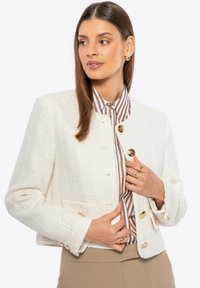 Giacca in tweed bianco con bottoni dorati, caratterizzata da un taglio corto e tessuto texturizzato. Abbinata a una blusa a righe marrone e bianca.