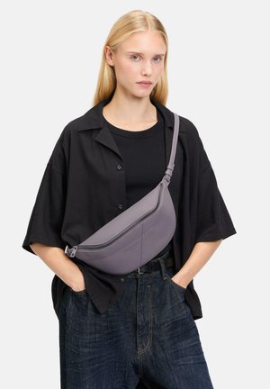Graue Leder-Umhängetasche mit kurvenförmigem Design und Reißverschluss, sichtbaren Nähten und einem verstellbaren Träger, der über einem schwarzen Oberteil getragen wird.