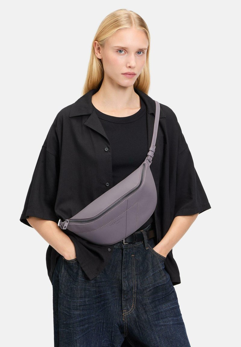 Grijze leren schoudertas met een gebogen vorm en een ritsluiting, voorzien van zichtbare stiksels en een verstelbare strap die gedragen wordt over een zwarte top.