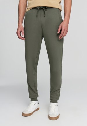 Pantaloni jogger verde scuro in morbido tessuto, con vita elastica e coulisse, gambe affusolate e polsini a coste. Indossati con sneaker bianche.