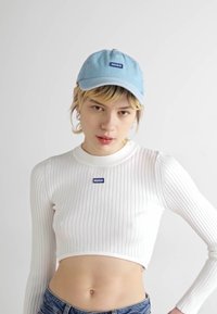 Lichtblauwe pet met "HUGO" logo, ribbelige witte crop top en high-waisted denim jeans. De top heeft een fitted ontwerp en ronde hals.
