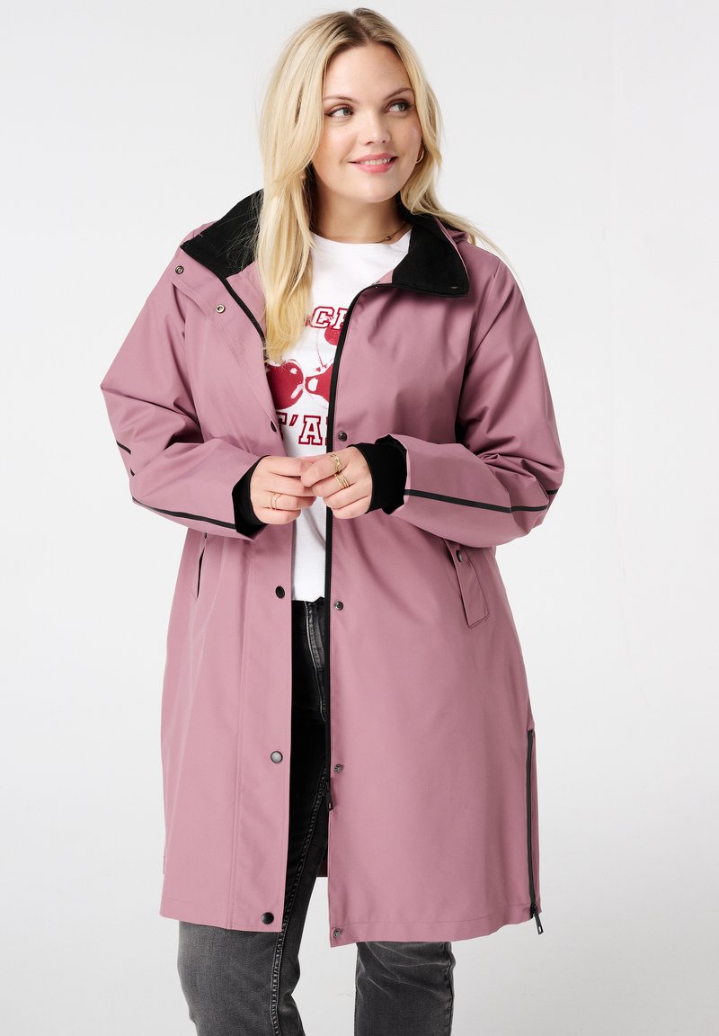 Blonde vrouw in een mauve lange jas over een witte overhemd met kersenprint en donkergrijze spijkerbroek, glimlachend en naar rechts kijkend.