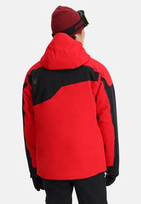 Rote und schwarze Skijacke mit Kapuze. Hat ein geometrisches Design, ein Logo auf dem Rücken und eine glatte Textur. Schwarze Akzente an den Ärmeln.