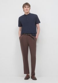 Giovane uomo con le mani in tasca, vestito con una camicia a maniche corte blu navy con colletto alla mandarino, pantaloni marroni, cintura intrecciata e mocassini marroni, in piedi di fronte a una parete bianca.