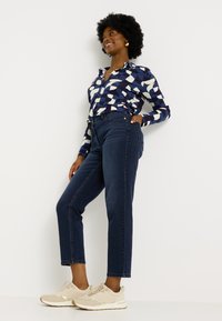 Chemise à motifs navy avec des formes géométriques, jeans sombres taille haute et baskets beiges. L'ensemble présente un col et une silhouette ajustée.