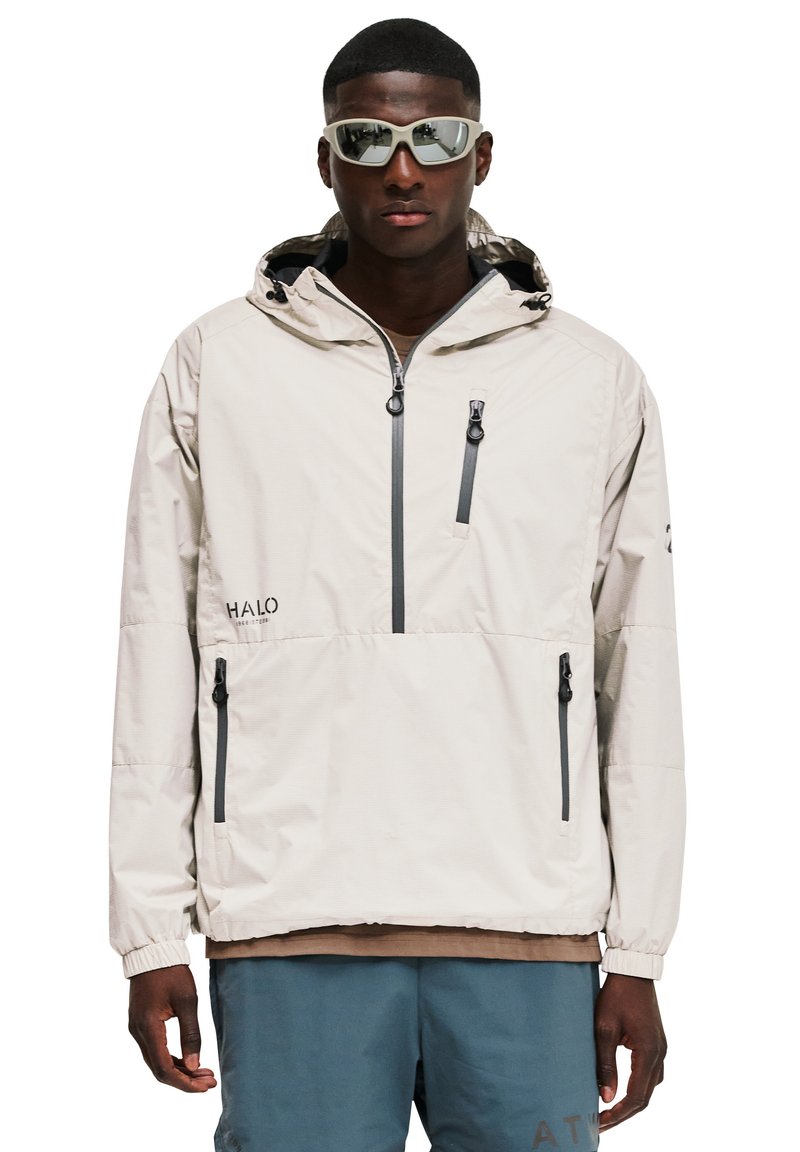 Mężczyzna ubrany w beżową kurtkę z kapturem, z przednimi zamkami błyskawicznymi i refleksyjnymi okularami przeciwsłonecznymi, stojący na białym tle.