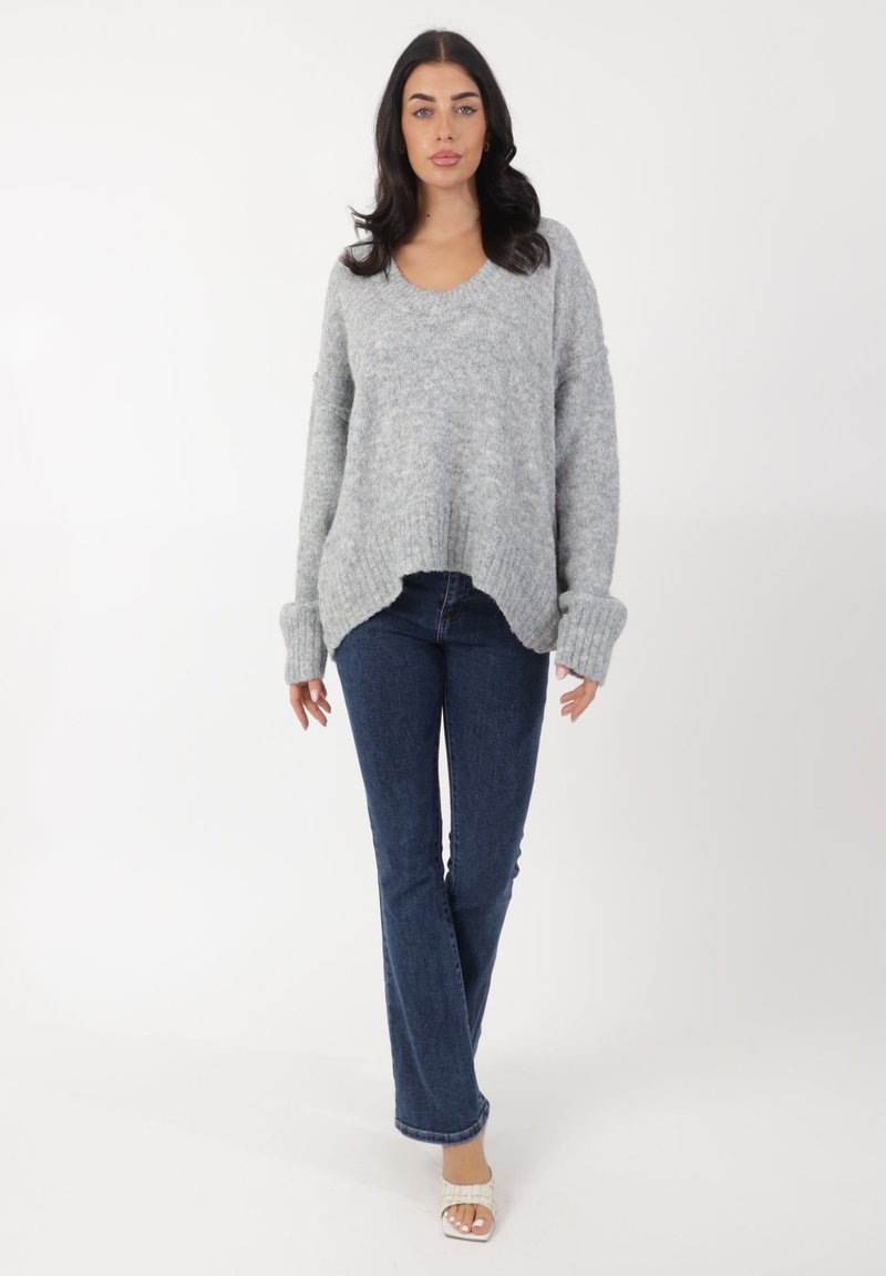 Grau gestrickter Pullover mit V-Ausschnitt und gerippten Bündchen, kombiniert mit blauen Schlagjeans. Die weiche Textur und der lockere Schnitt betonen den Komfort.
