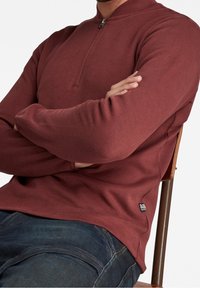 Pull en tricot bordeaux avec un col à quart de zip, des manches longues et des poignets côtelés. Comprend une petite étiquette de marque sur le côté gauche.