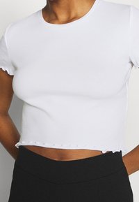 Biała krótka koszulka z rypsowej tkaniny, z okrągłym dekoltem i falbaniastymi brzegami rękawów oraz dołu, zestawiona z czarnymi spodniami o wysokim stanie.