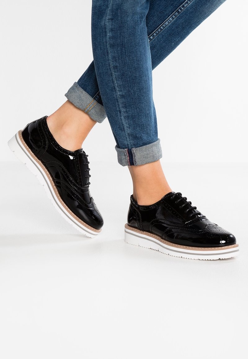 Personne portant des brogues en cuir verni noir avec des semelles blanches et un jean bleu retroussé marchant sur un sol blanc.