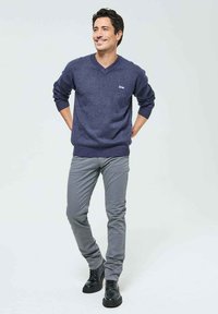 Pull en V bleu marine en tricot, associé à un pantalon slim gris clair et des chaussures noires à lacets. Présente un logo sur la poitrine.