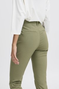 Femme portant un pantalon ajusté vert olive et un haut blanc à manches longues, debout avec une main reposant le long du corps.