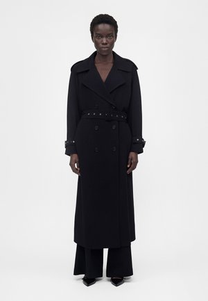 Femme debout face à l'objectif, portant un long trench-coat noir ceinturé, un pantalon noir et des chaussures pointues noires sur un fond uni.