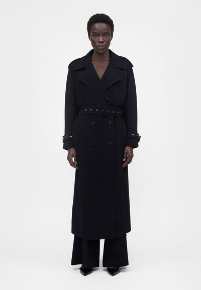 Femme debout face à l'objectif, portant un long trench-coat noir ceinturé, un pantalon noir et des chaussures pointues noires sur un fond uni.