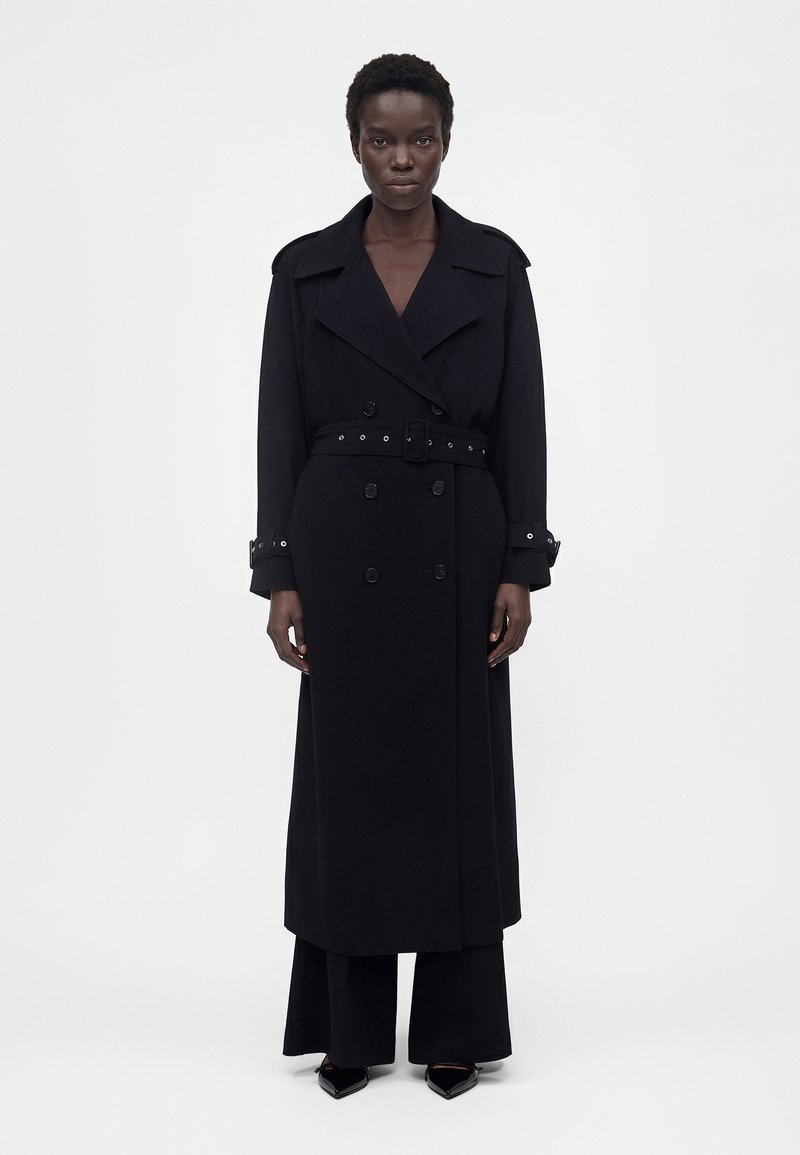 Γυναίκα που στέκεται μπροστά προς την κάμερα, φορώντας μακρύ μαύρο παλτό με ζώνη, μαύρο παντελόνι και μαύρα μυτερά παπούτσια, σε απλό φόντο.