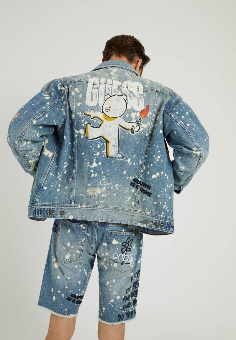 Jeansjacka och shorts-set med stänkmönster, tecknad björn-illustration och "GUESS"-text, i ljusblått med slitna detaljer.