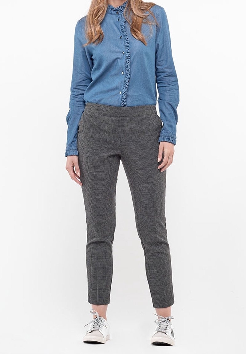 Chemise en denim bleu clair avec devant à volants, associée à un pantalon slim à carreaux gris. Le mannequin porte des baskets blanches.