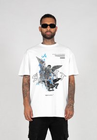 Hvid bomulds T-shirt med en grafisk print af en vinget figur med blå accenter og tekst. Løs pasform med korte ærmer. Sorte cargo shorts.