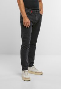 Jeans in denim nero con una vestibilità slim, caratterizzati da un accento rosso nella cintura e dal classico design a cinque tasche, abbinati a sneakers bianche.