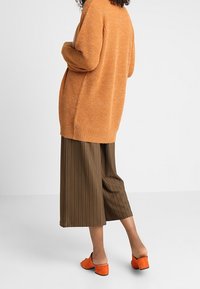 Cárdigan naranja de gran tamaño con canalé, amplios culottes marrones con pliegues y mules naranjas con tacón bloque. Textura suave, corte relajado y colores vibrantes.