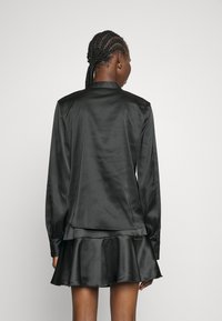 Blusa nera in satin con maniche lunghe, colletto strutturato e retro leggermente drappeggiato. Completata con polsini abbottonati e una texture liscia.