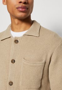 Cardigan en tricot beige avec un motif texturé, col, fermeture à boutons sur le devant et poche poitrine. Présente des boutons marron sur le devant.