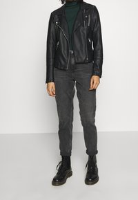 Schwarze Lederbikerjacke mit Reißverschlüssen, grüner strukturiertes Sweatshirt, graue Slim-Fit-Jeans und schwarze Schnürstiefel.