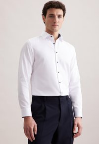 Man in een aansluitend wit overhemd met knopen en donkere formele broek, staand tegen een effen lichte achtergrond.