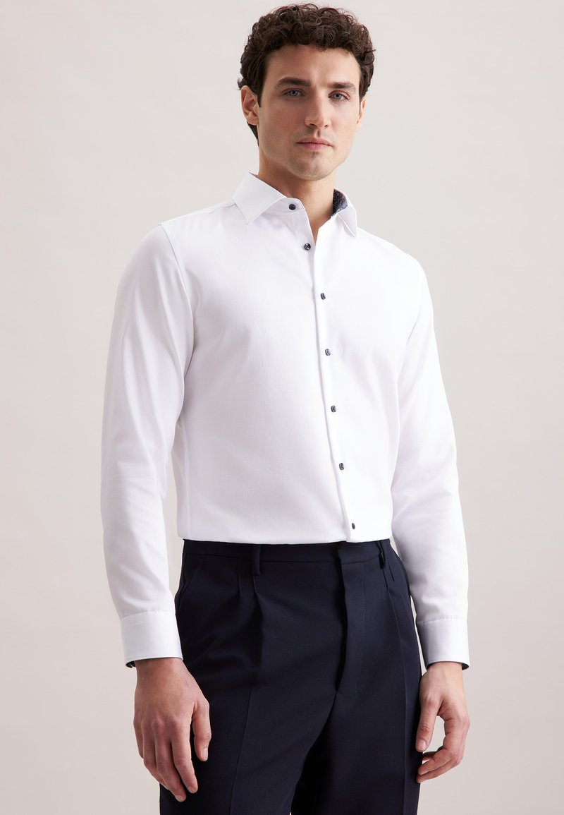 Man in een aansluitend wit overhemd met knopen en donkere formele broek, staand tegen een effen lichte achtergrond.