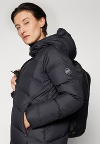 Svart puffjacka med hög krage och huva, med en zippered ficka och en logotyp på ärmen. Buren över en svart ryggsäck.