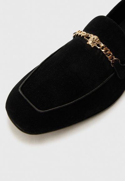 Mocassins en daim noir avec un orteil carré, dotés d'une accent en chaîne dorée et d'un détail de tête de lion. Texture lisse et coutures soulignées.
