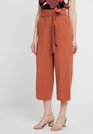 Kvinde iført rustfarvede cropped bukser med vidde ben og bindebælte i taljen samt beige blokhælede sandaler, stående mod en ensfarvet baggrund.