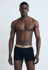 Schwarze Boxershorts mit einem goldenen "BOSS"-Logo am Bund, aus glattem Material gefertigt. Das Modell zeigt eine körpernahe Passform.