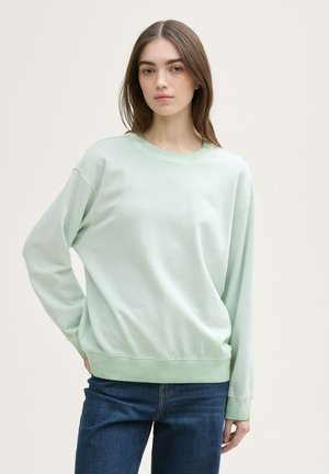Hellgrüner Sweatshirt mit Rundhalsausschnitt und fallenden Schultern. Weiches Baumwollmaterial und gerippte Bündchen. Kombiniert mit blauen Jeans.