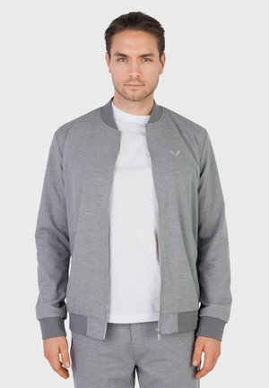 Uomo che indossa una giacca grigia chiaro con cerniera sopra una camicia bianca e pantaloni grigi coordinati, rivolto verso la fotocamera su sfondo semplice.