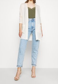 Light beige cardigan with long sleeves and front pockets, layered over an olive green camisole, paired with high-waisted light blue jeans.