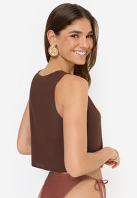 Καφέ κοντό τοπ από ελαφρύ ύφασμα με στρογγυλή λαιμόκοψη. Το μοντέλο φοράει χρυσα αυτιών σπείρας, παρουσιάζοντας μια λεία υφή.