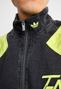 Svart jeansjacka med hög krage, limegröna detaljer och logotyp. Har en metallkedja och texturerat tyg.