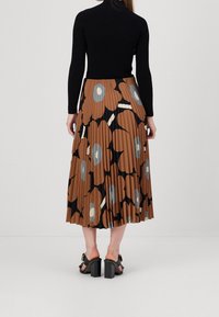 Donna che indossa un top nero a maniche lunghe, una gonna midi plissettata marrone con una grande stampa floreale e sandali neri con tacco, in piedi su uno sfondo bianco.