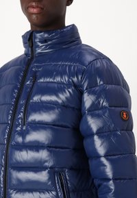 Marineblå dunjakke med en blank finish, lynlåslukning foran, høj krave og et mærkedetalje på venstre arm. Har vandrette syninger.