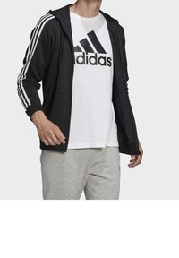 Persona che indossa una felpa nera con zip e strisce bianche sulle maniche, una maglietta bianca Adidas e pantaloni della tuta grigio chiaro.