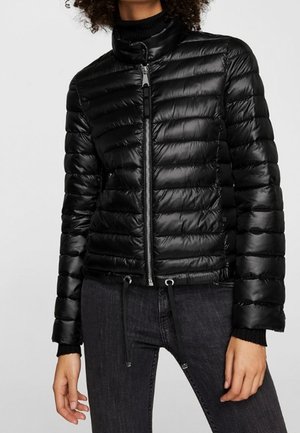 Osoba ubrana w czarną pikowaną kurtkę puchową z przednim zamkiem błyskawicznym i ściągaczem, w połączeniu z dopasowanymi ciemnoszarymi jeansami.