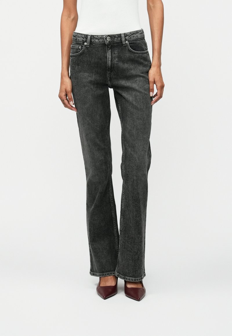 Jeans en denim noir avec une coupe droite, des accents cousus, deux poches avant et une fermeture à bouton classique. Portés avec des talons aiguilles bordeaux.