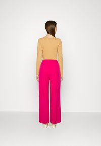Top beige a maniche lunghe abbinato a pantaloni rosa a vita alta e gamba larga, che mostrano una texture liscia e una vita aderente, visto da dietro.