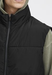 Schwarze Steppweste mit hohem Kragen und Reißverschluss, mit strukturiertem Oberflächenmaterial. Wird über einem hellgrünen Hoodie getragen.